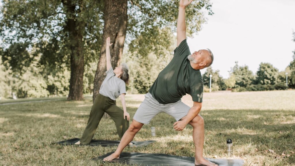 A man and woman in yoga poses outside near trees.