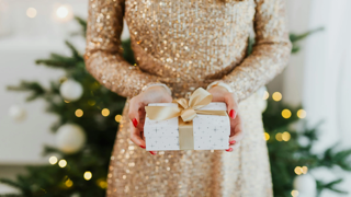 Woman standing in front of a lit Christmas tree, holding a wrapped gift.